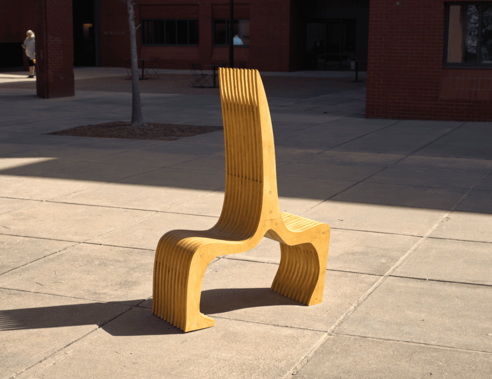 Installed piece photographed alone in Dudley Courtyard, full form visible — high curved back, splayed legs, and laminated plywood slats casting sharp shadows across pavement.
