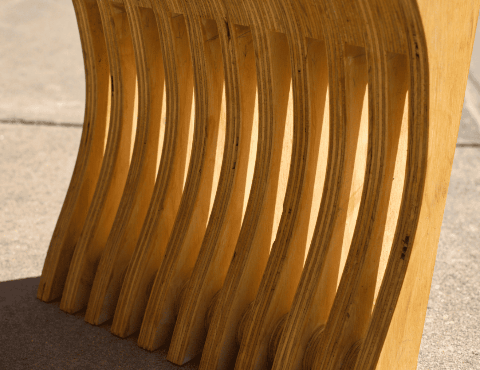 Close detail of the laminated plywood base reveals layered grain depth, CNC-cut curvature, and the rhythmic slit pattern running through each structural member.