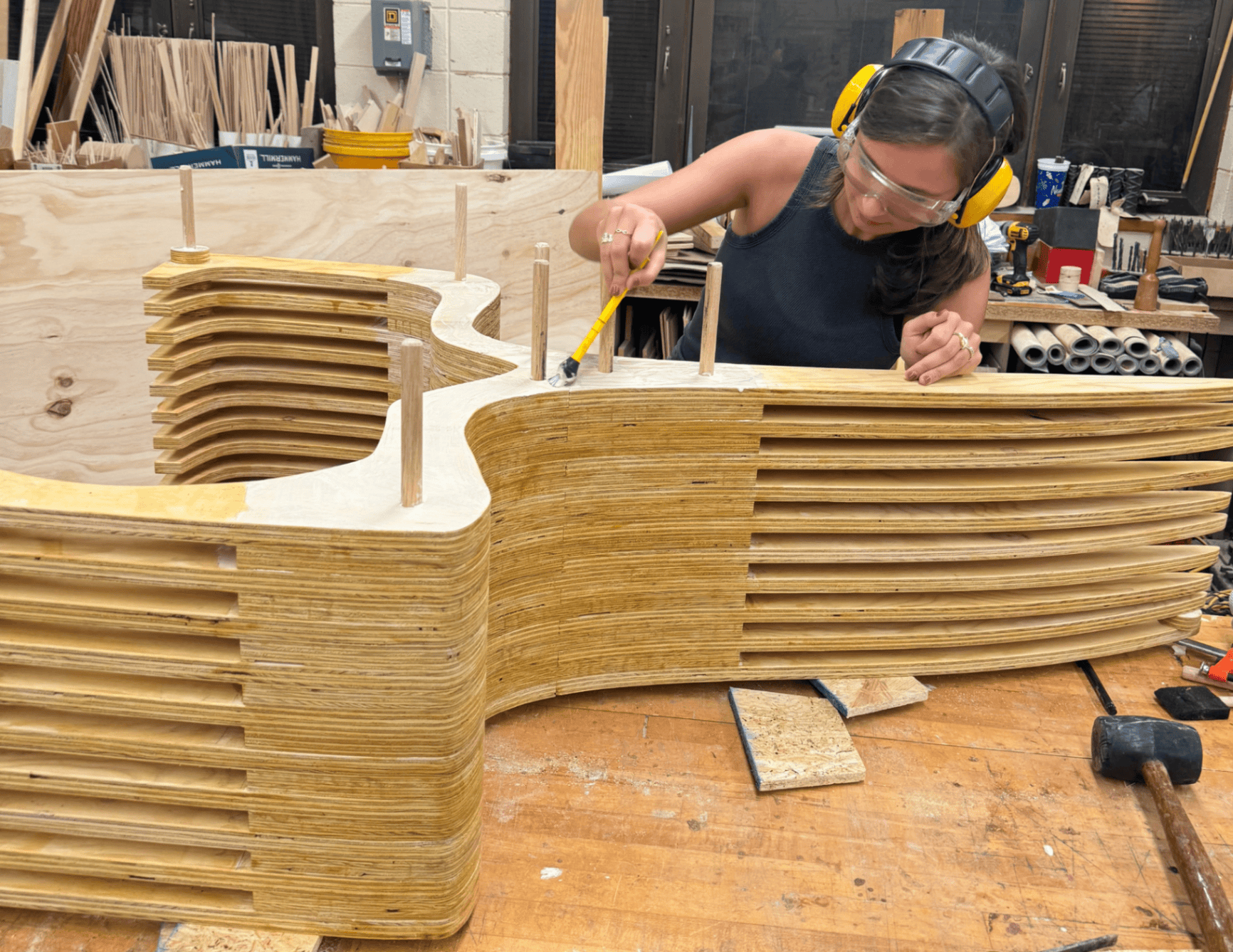 Annie assembles stacked plywood layers at the workbench, applying glue between CNC-cut sections with dowel registration pins holding alignment during lamination.