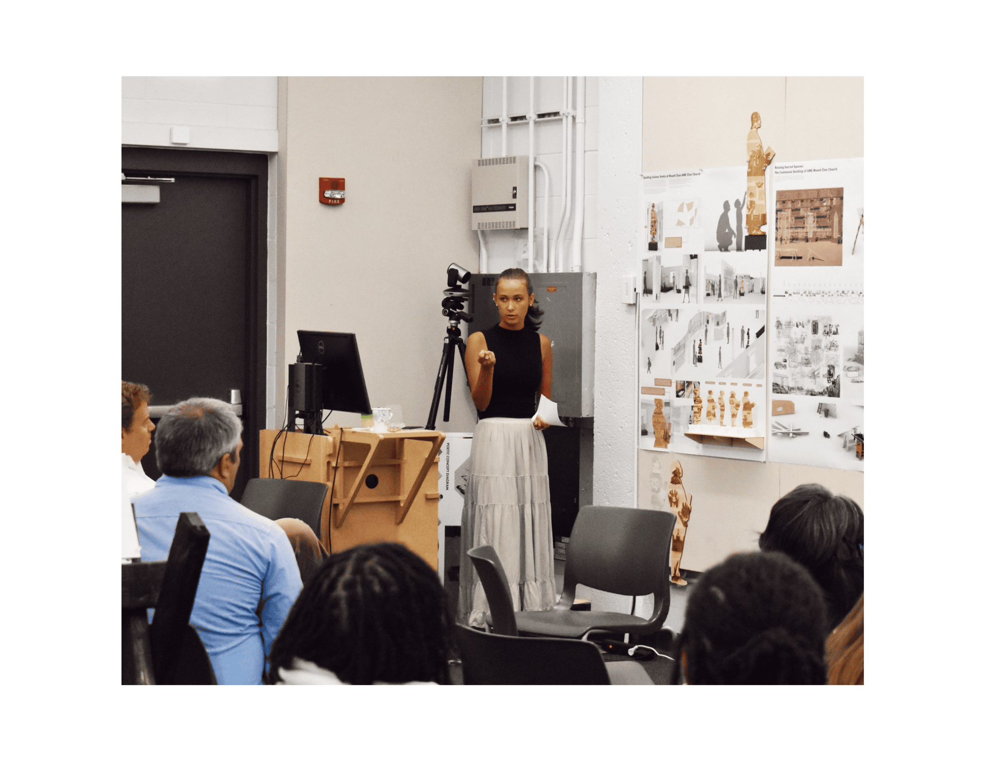 Annie presents the project to church community members and restoration architects, with fabricated figures and full presentation boards visible behind her.