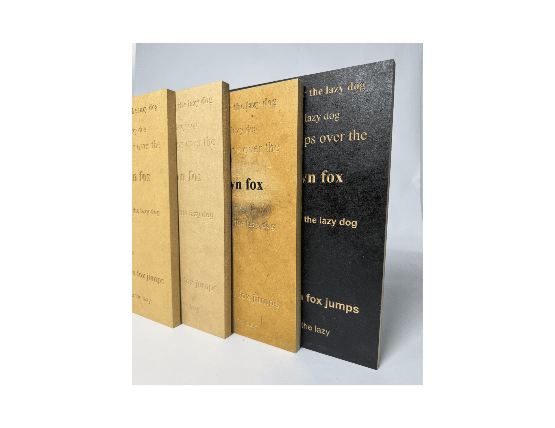Four MDF material samples display CNC-engraved text at varying stain depths and finish tones — from raw natural to deep matte black — testing legibility and surface contrast.