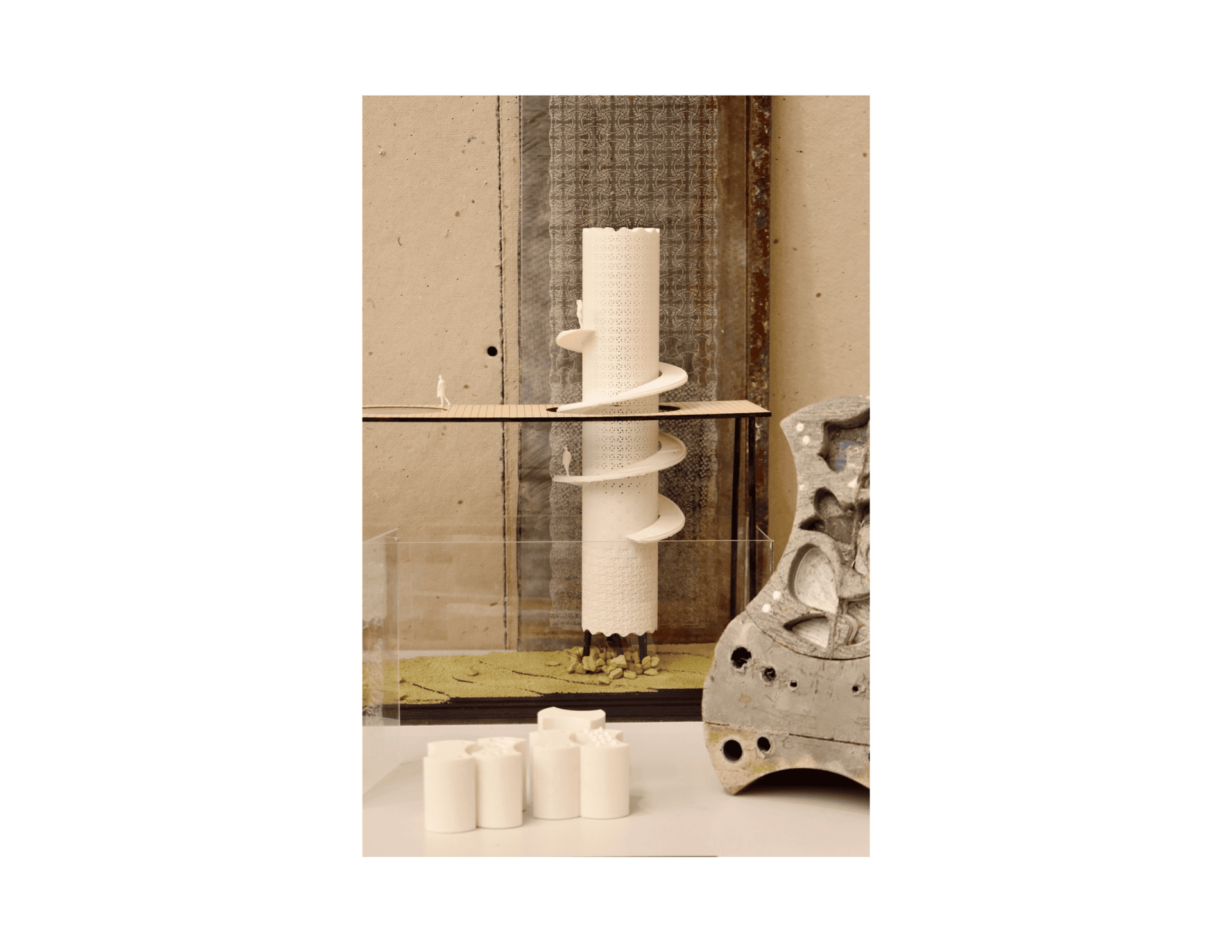 Physical model tableau groups the 3D-printed perforated tower, module prototypes, and concrete ecology cast against rusted metal and etched acrylic backdrop panels.